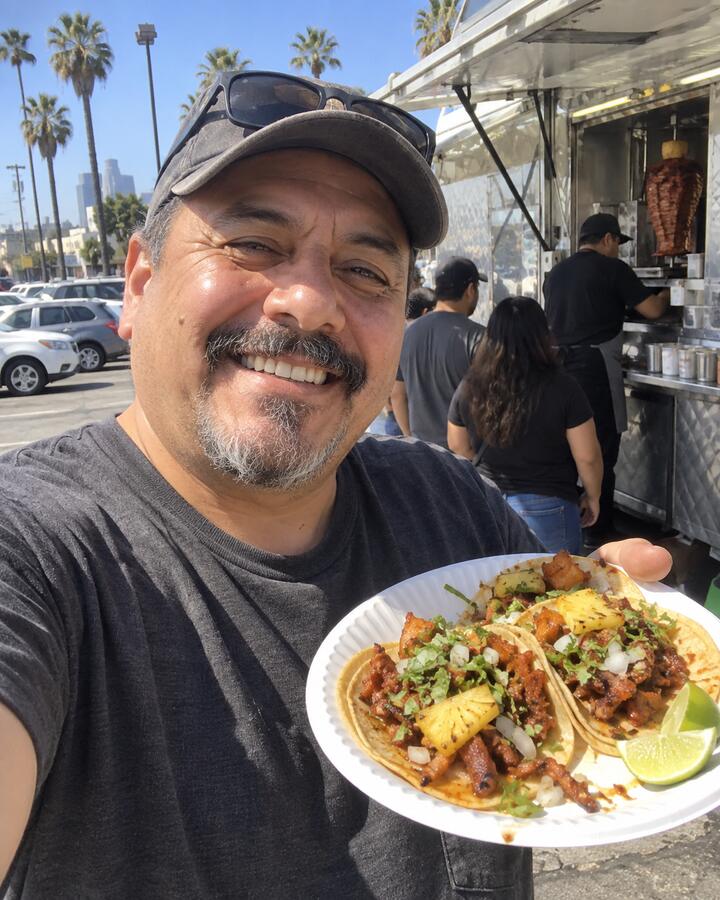 A man taking a selfie at a taco truck holding a plate of tacos